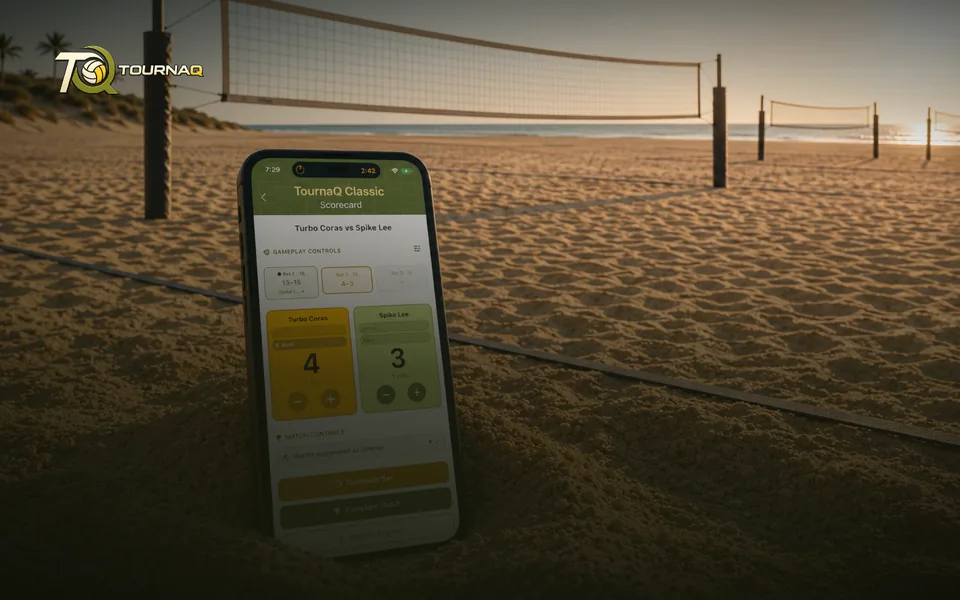 A phone standing in the sand with a live TournaQ scorecard, beach volleyball courts behind it at sunset